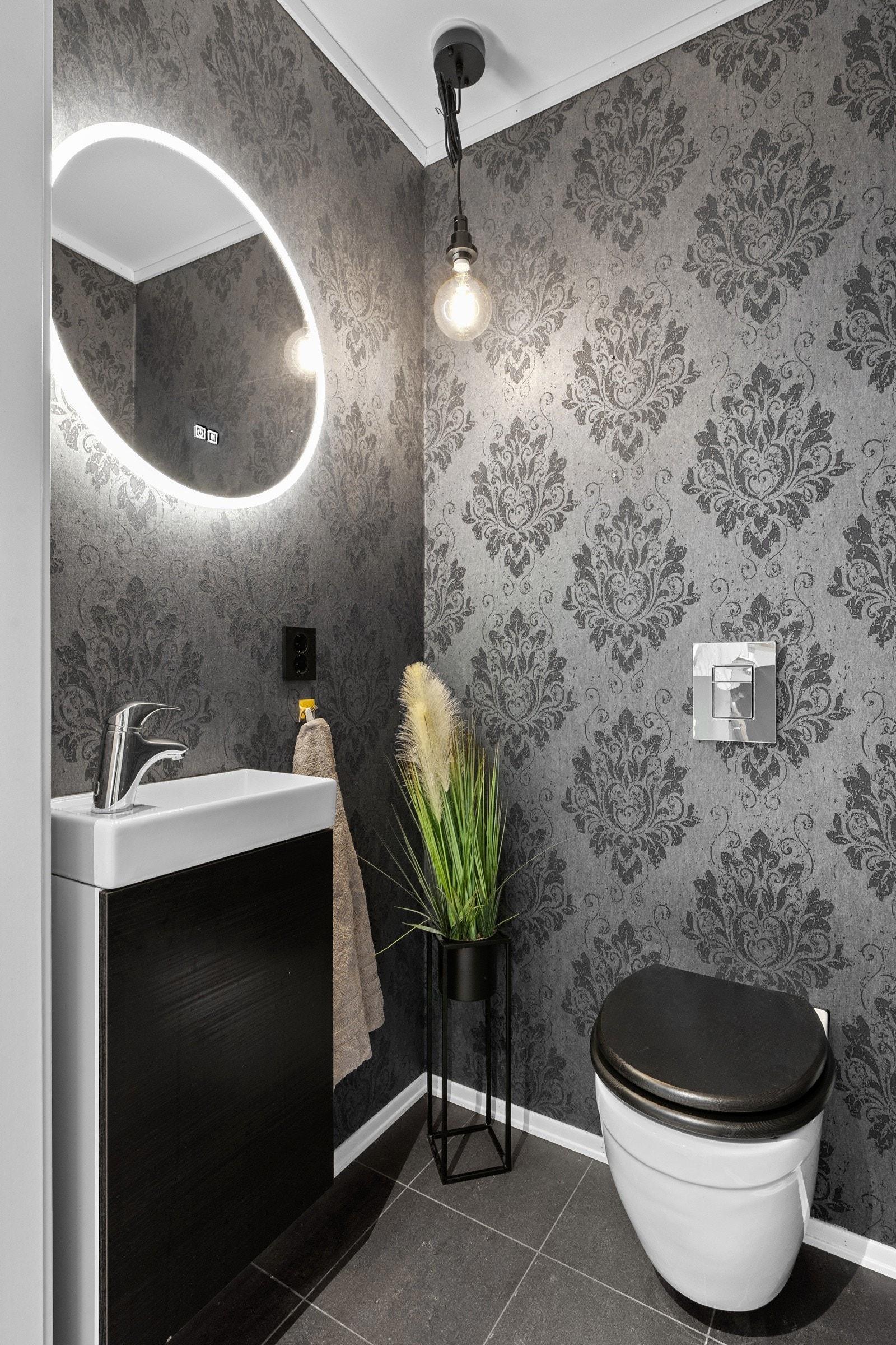 Praktisk, seperat toalettrom med tapetserte vegger og flislagt gulv med gulvvarme. Rommet inneholder en servant med underskap, vegghengt toalett og speil. Galleribilde