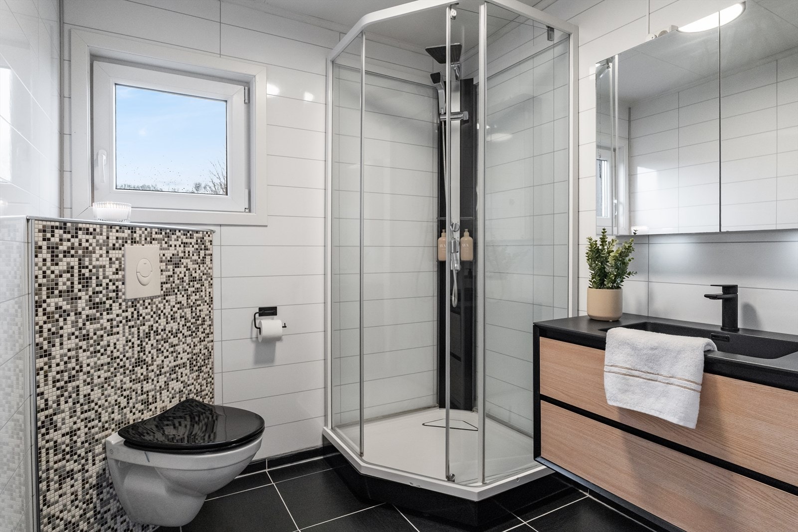 Pent bad med flislagt gulv med varmekabler og baderomsplater på veggene. Badet er innredet med vegghengt toalett, dusjkabinett og en nyere baderomsinnredning med servant og speil. Galleribilde