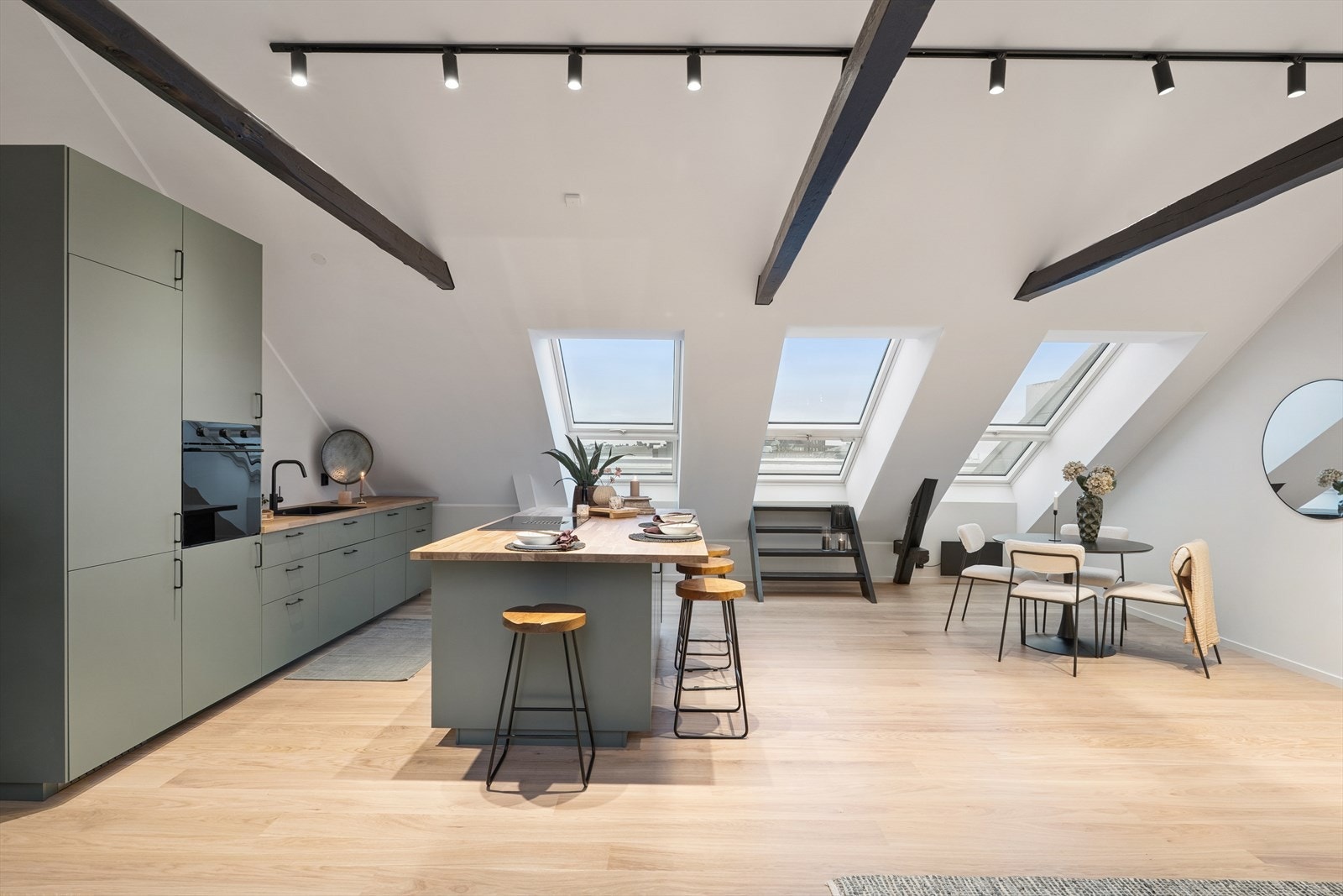 Stue og kjøkken ligger i åpen løsning og har lysmalte flater, lys enstavs parkett på gulv, store takvindu som gir rikelig med naturlig lys og synlige, sortmalte takbjelker som brekker av det lyse og setter et stilig preg på leiligheten. Galleribilde