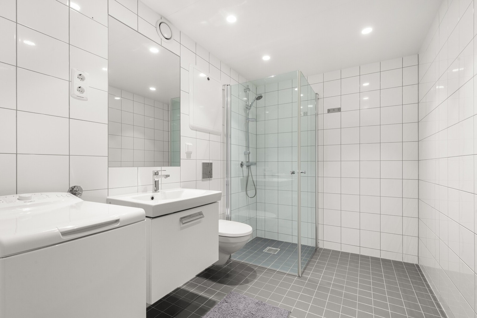 Dusj med innfellbare glassvegger, servantseksjon med skuff og heldekkende porselenvask, innmurt speil, vegghengt wc, opplegg for vaskemaskin og tørketrommel. Galleribilde