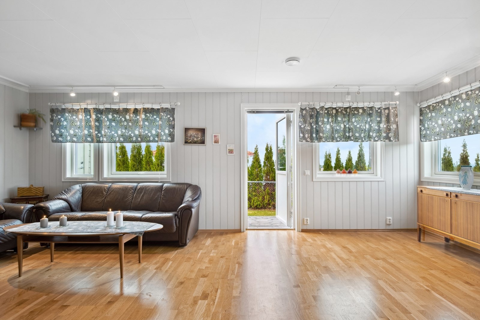 Lys og trivelig stue med parkett på gulv, panelplater i en dus, lysegrå fargetone på veggene, og takessplater i himlinger, som er gjennomgående i hele boligen. Galleribilde