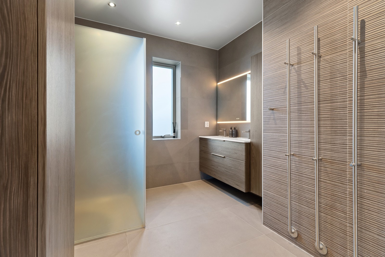 Smakfullt hovedbad i boligens 2. etasje med delikate storformatfliser på gulv- og veggflater, samt varmekabler i gulv. Innredningen består av stilrent servantmøblement med 2 skuffer, speil, 2 stk. tilhørende høyskap og vegghengt toalett. Galleribilde