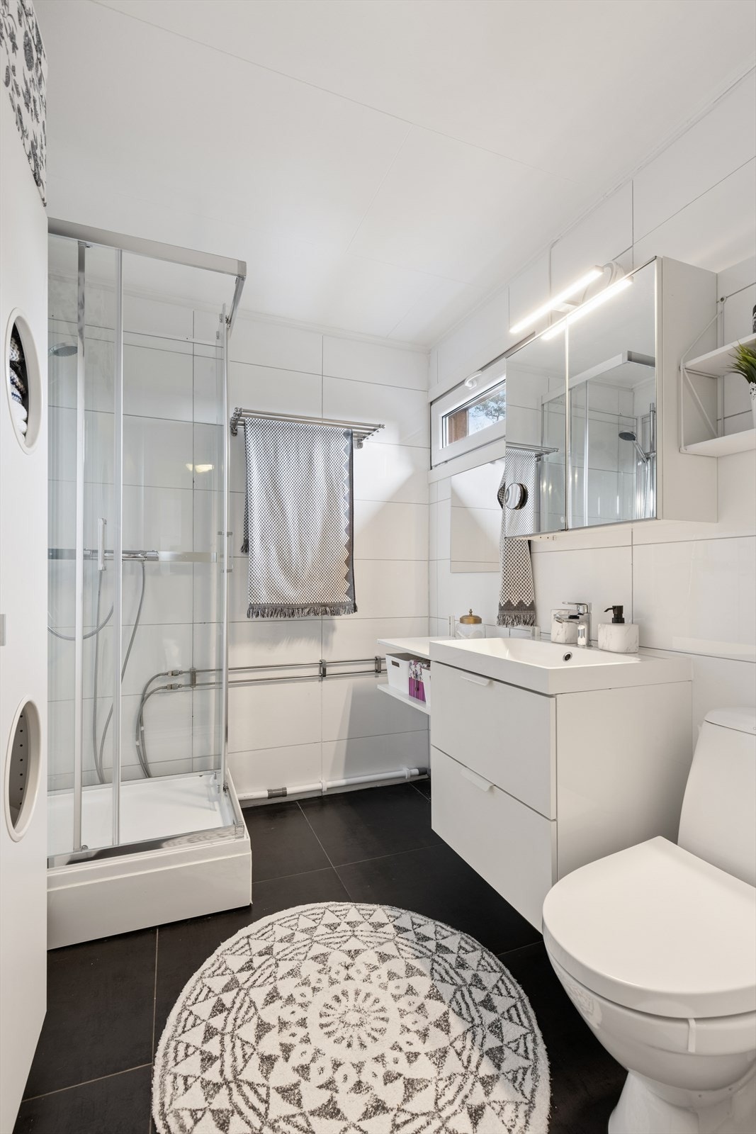 Badet har fliser på gulv og vegger, utstyrt med bla. dusjkabinett, toalett, servant og speilskap med belysning. Galleribilde