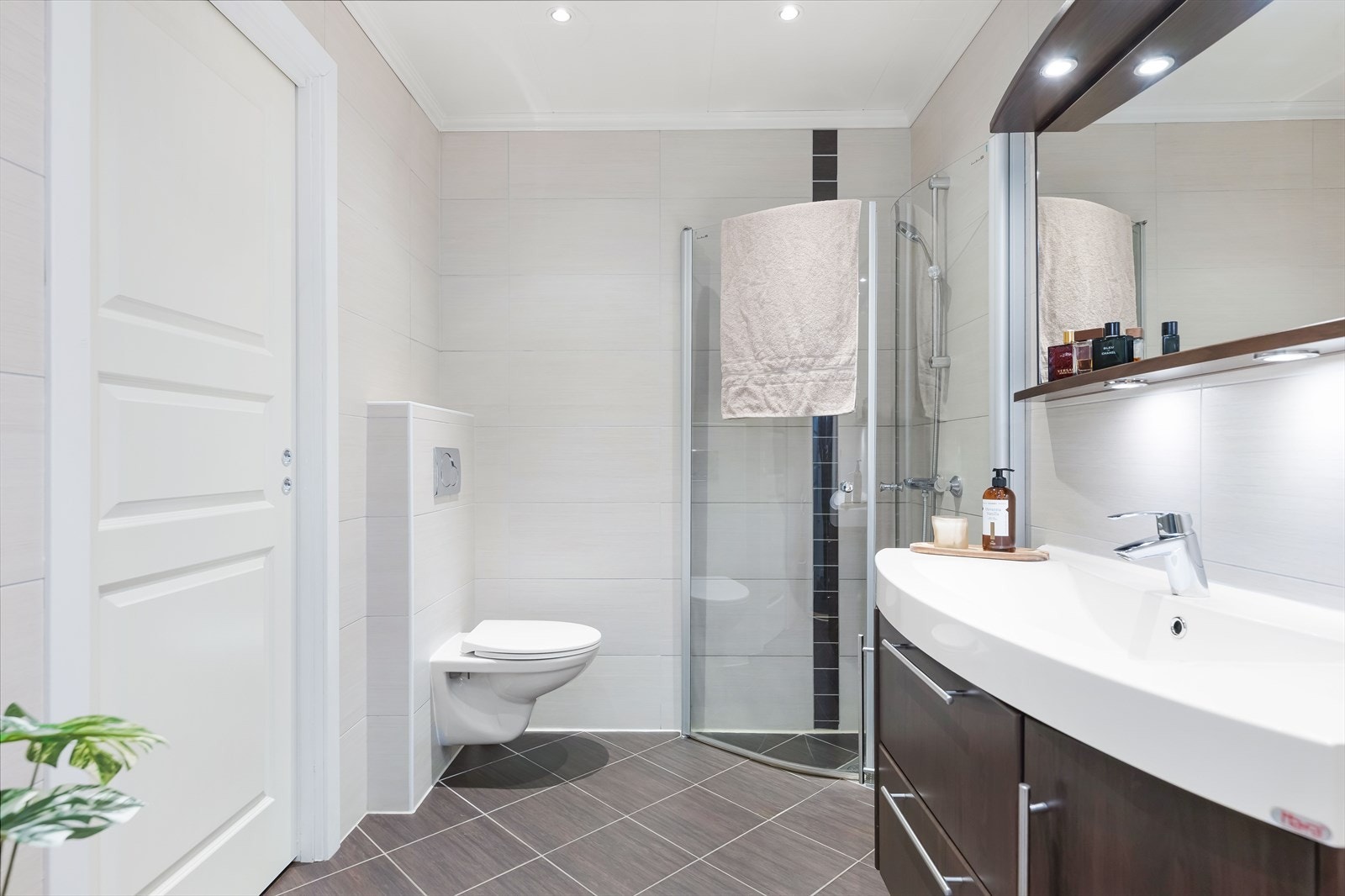 Stilrent bad med benkeskap med nedfelt servant og speil med belysning over. Badet er utstyrt med vegghengt toalett og dusjhjørne. Galleribilde