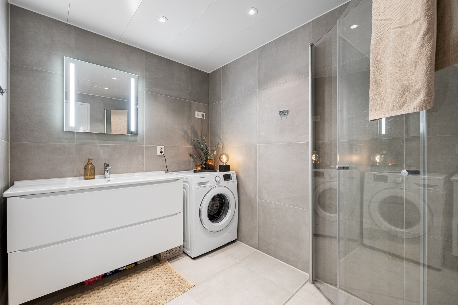 Baderommet er innredet med innfellbare og plassbesparende dusjdører i glass, dusjbatteri og garnityr montert på vegg, kran og avløp for vaskemaskin og veggmontert WC. Galleribilde