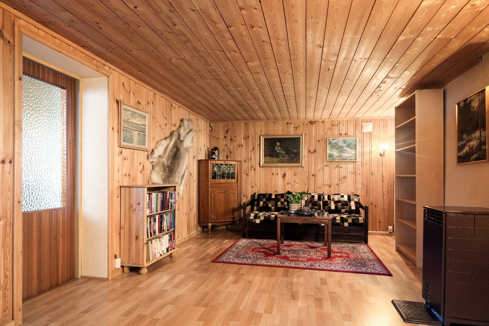 Veggene er kledd i klassisk furupanel, og taket følger opp med panel som gir rommet en lun og helhetlig ramme. Laminatgulvet gjør det lettstelt og anvendelig, mens vedovnen sørger for ekte varme og stemning når kulden setter inn. Galleribilde