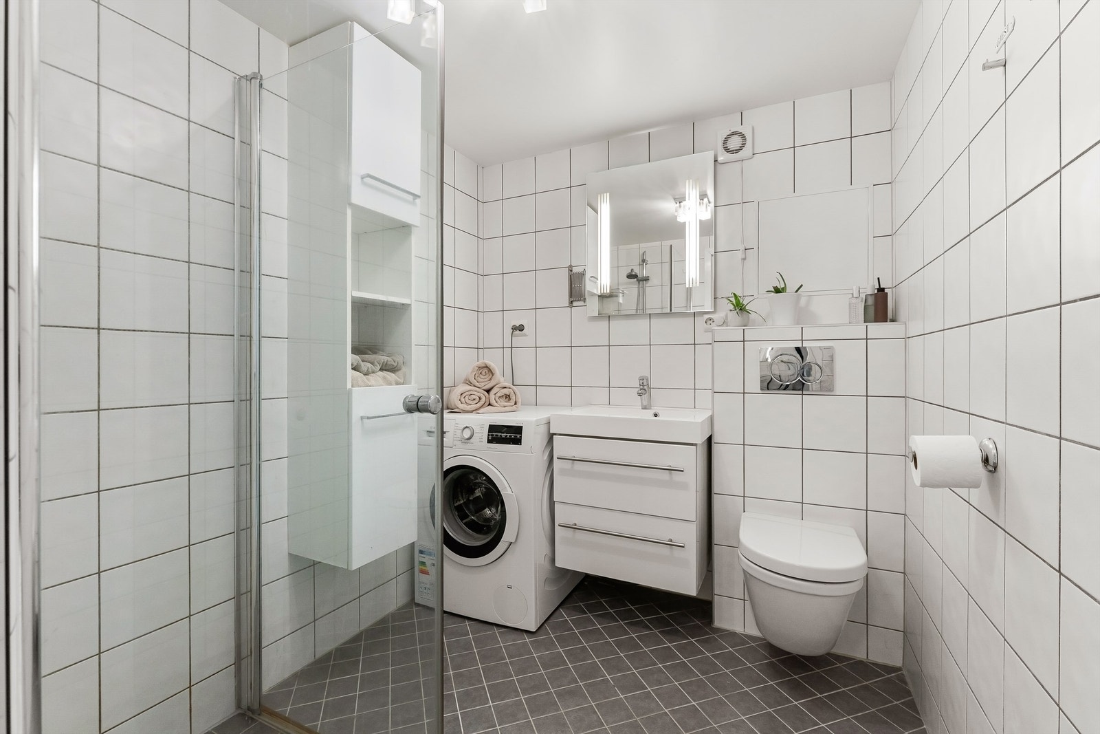 Flislagt bad med vegghengt toalett, dusjhjørne og opplegg for vaskemaskin. Galleribilde