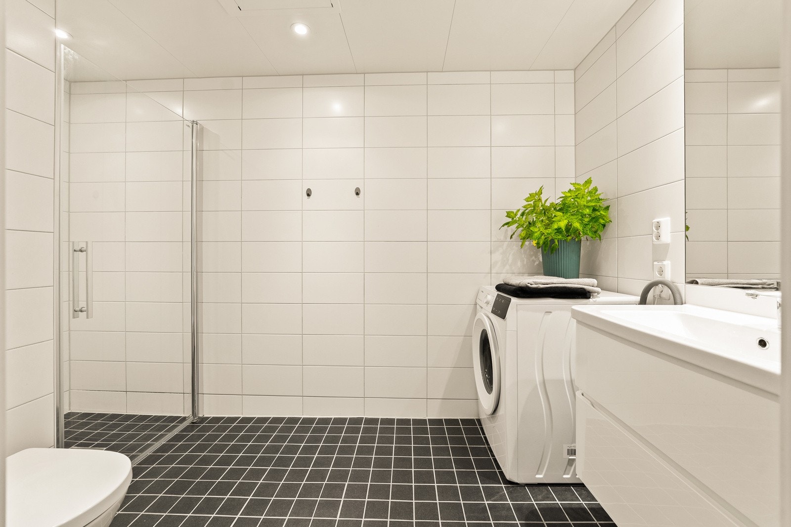 Bad. Her er det hvite fliser på veggene, downlights i himling og mørke gulvfliser med varme. Servant, veggfestet klosett med innebygget sisterne, dusj med innfellbare dør i herdet glass og opplegg for vaskemaskin. Galleribilde