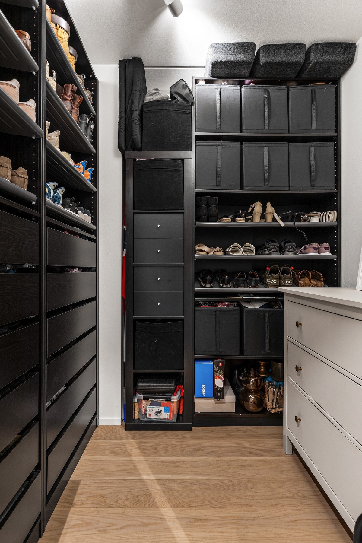 I tilknytning til entréen ligger en bod innredet med sorte, åpne skap med skuffer, hyller og opphengsstenger for oppbevaring av yttertøy og sko Galleribilde