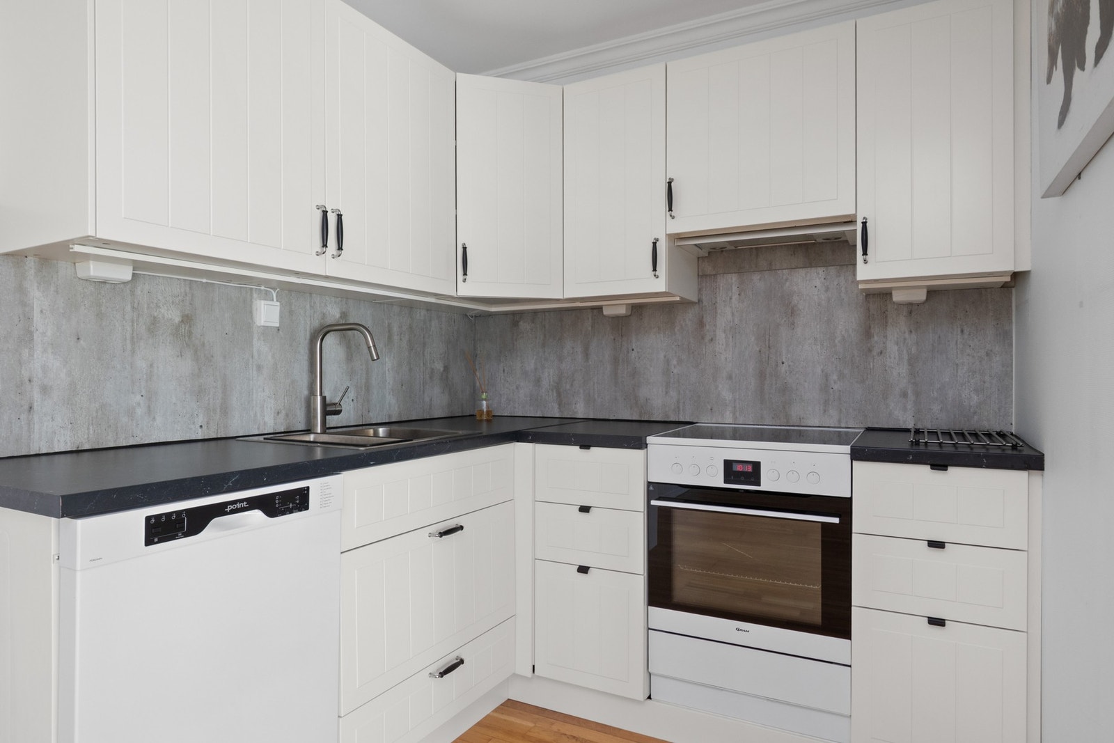 Innredningen er fra 2025, oppført i en liten L-form med fin skapplass, hvite, profilerte fronter, dobbel oppvaskkum og sort benkeplate av laminat Galleribilde