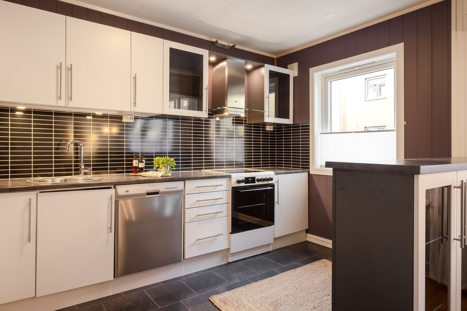 Innredningen har hvite glatte fronter og laminert benkeplate med nedfelt oppvaskkum. Praktisk med flisimitasjonsplater i benkeryggen og belysning under overskapene. Galleribilde