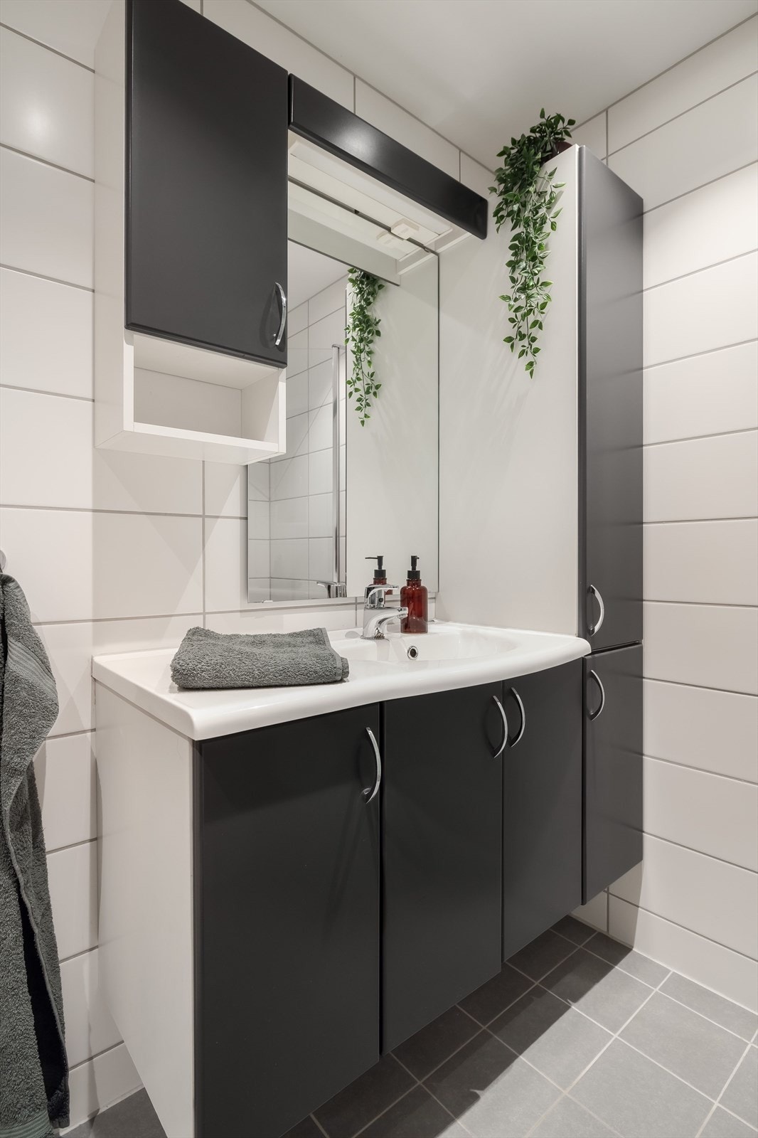 Tidløs baderomsinnredning med mørke glatte fronter og heldekkende servant, speil med belysning og vegghengt skap over. Galleribilde