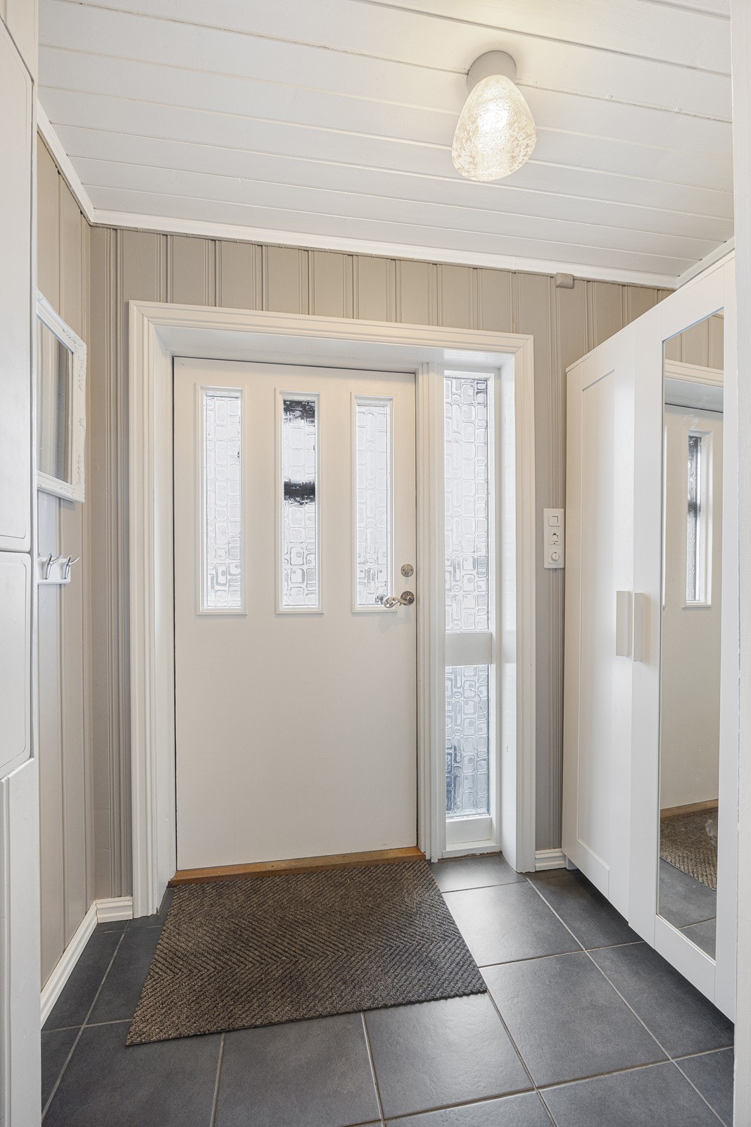 Vel inne blir vi ønsket velkommen i et pent vindfang med gråmalt panel på veggene og grå fliser på gulv. Galleribilde