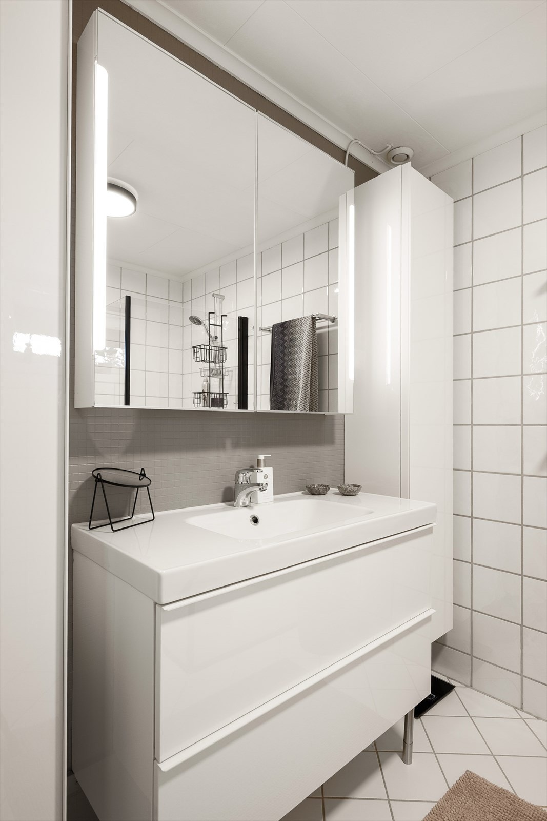 Badet er innredet med hvit baderomsinnredning med heldekkende servantplate, speilskap med integrert belysning, veggmontert toalett og dusjhjørne med innfellbare dører. Galleribilde