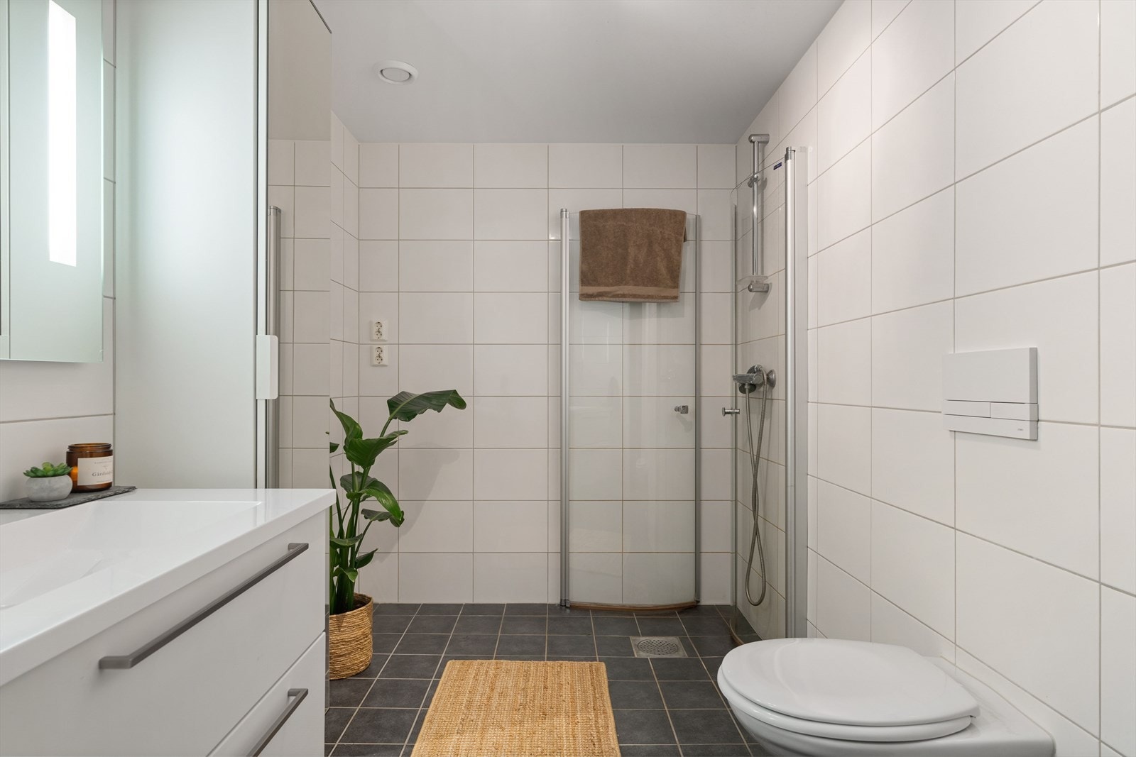 Badet er innredet med servant i moderne underskap, speil med integrert belysning, vegghengt toalett og dusjhjørne med glassdører. Galleribilde