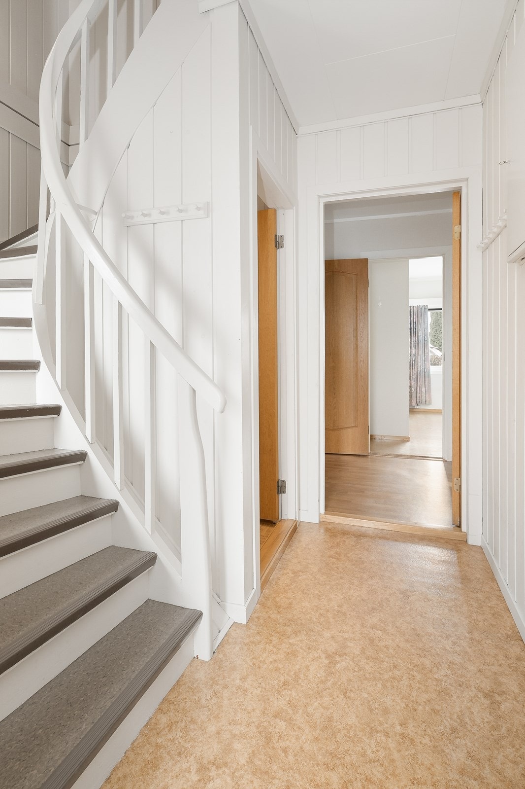 Via inngangsdør fra 2015 kommer man inn i boligens vindfang med tilliggende trappegang med lysmalte veggoverflater. I trappegangen er sikringsskapet plassert. Galleribilde