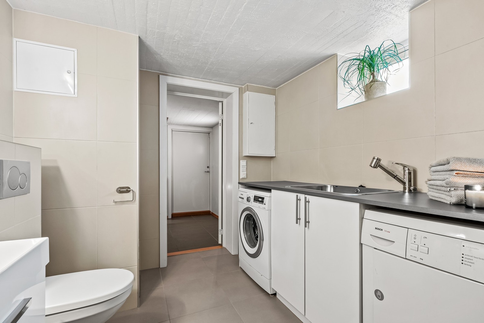 På motsatt vegg er det skap under en laminatbenkeplate med nedfelt kum i stål. Under benken er det opplegg og plass for både vaskemaskin og tørketrommel. Galleribilde