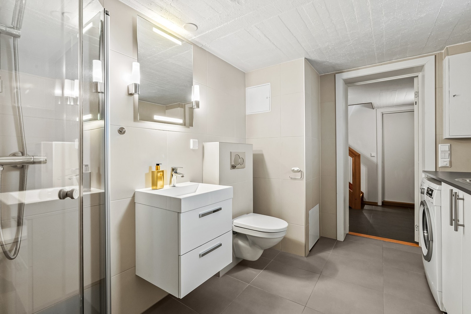 Badet er utstyrt med et vegghengt toalett og et dusjhjørne med innfellbare glassvegger. Baderomsinnredningen består av en heldekkende servant på en skuffeseksjon og et speil med belysning. Galleribilde