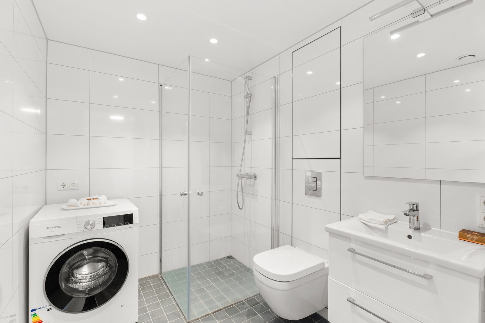 Meget pent flislagt bad med gulvvarme. Badet har dusjhjørne med dører i herdet glass, veggfestet klosett, servant og opplegg for vaskemaskin/tørketrommel. Galleribilde