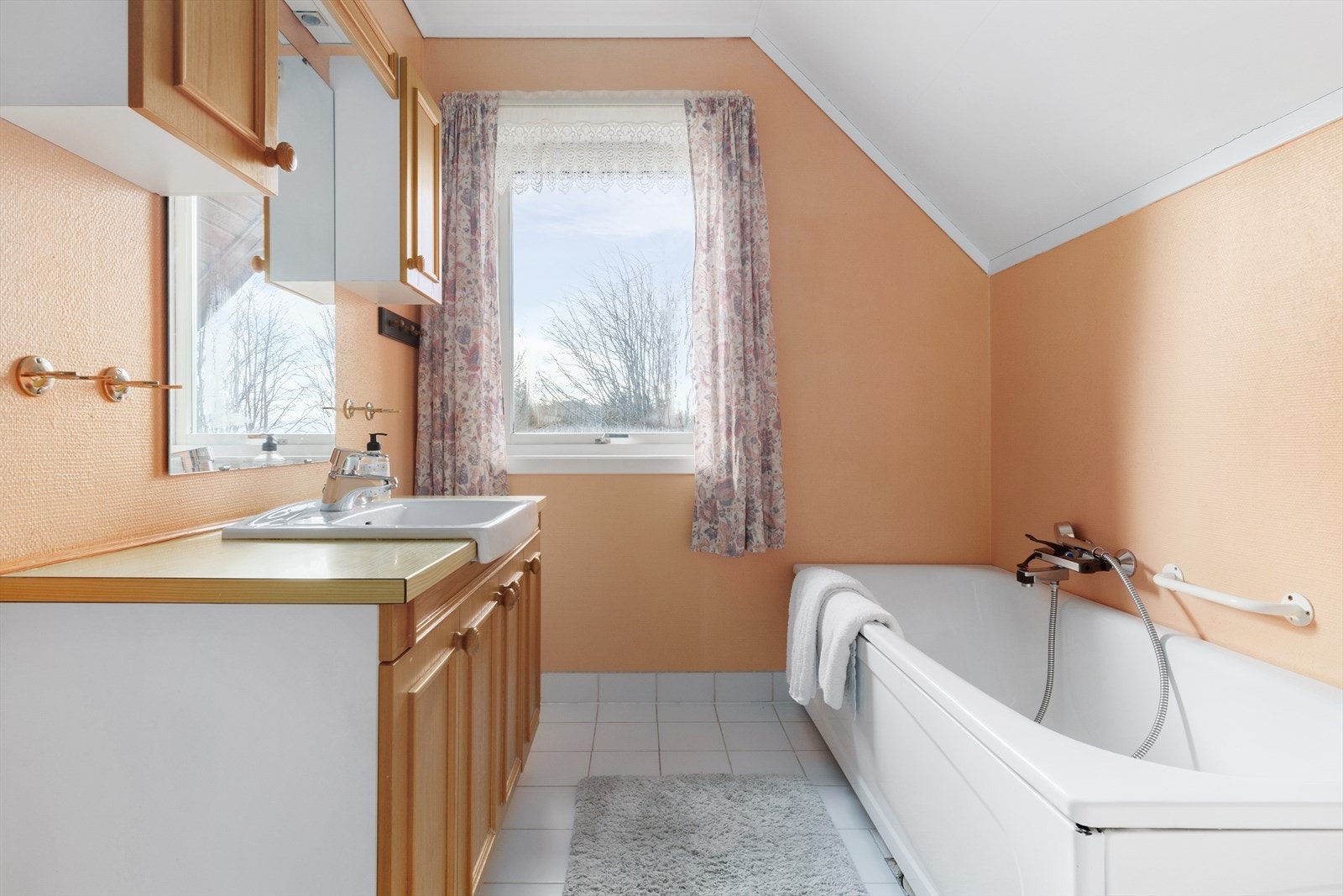 Badet er laget med fliser på gulv og tapet på veggene. Badekar, servant med innredning og overskap. Galleribilde