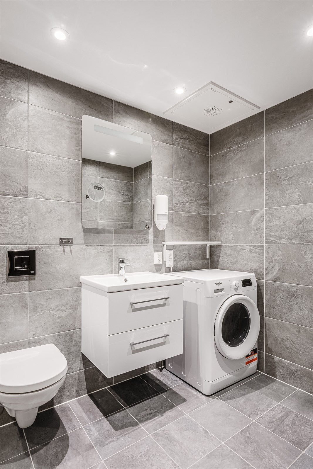 Moderne baderomsinnredning med skuffer og heldekkende servant, speil med integrert belysning over. Det er også plass og opplegg for vaskemaskin. Galleribilde