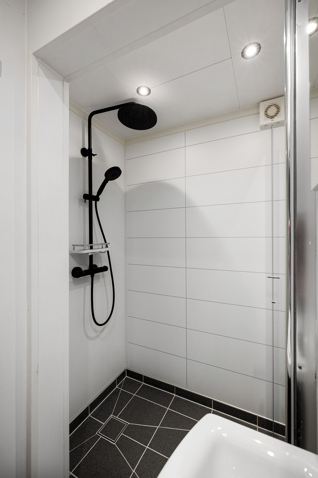 Badet har fliser på gulv og baderomsplater på vegger. Innredningen består av et underskap/overskap, servant, speil, lys, stikk og en dusjsone med dusjing direkte på gulv og vegg. Galleribilde