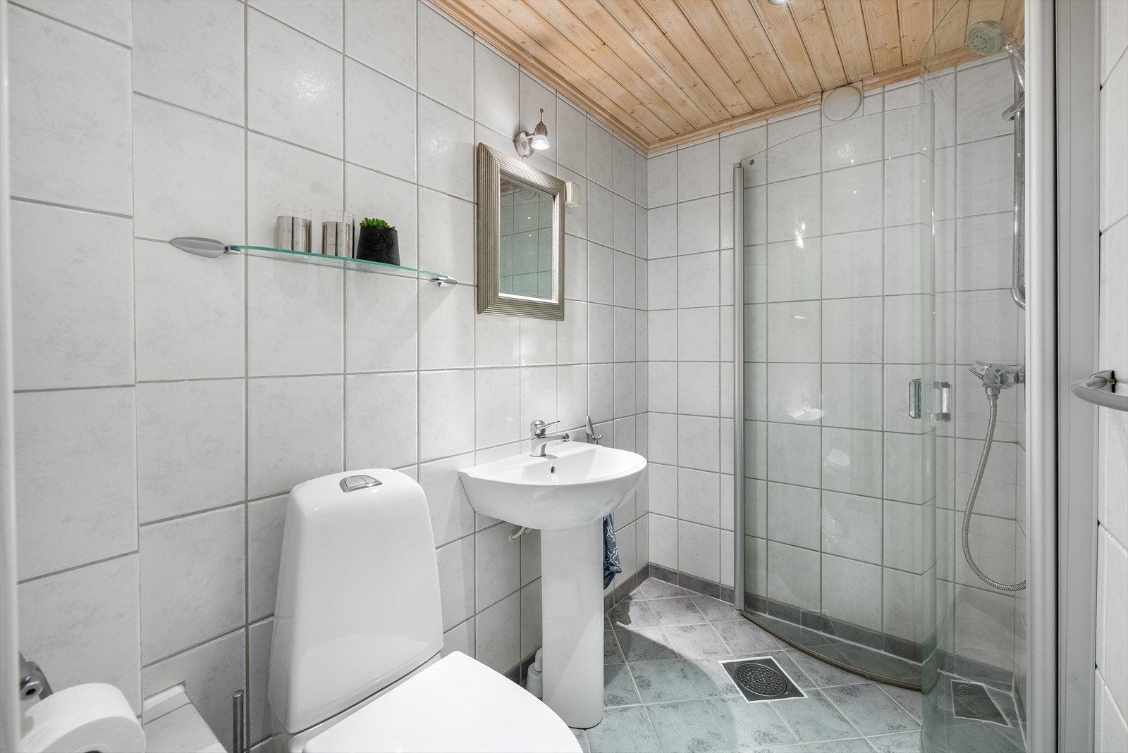 Pent flislagt baderom i lyse fargetoner. Badet er utstyrt med toalett, servant og dusjnisje med innfellbare glassdører. Galleribilde