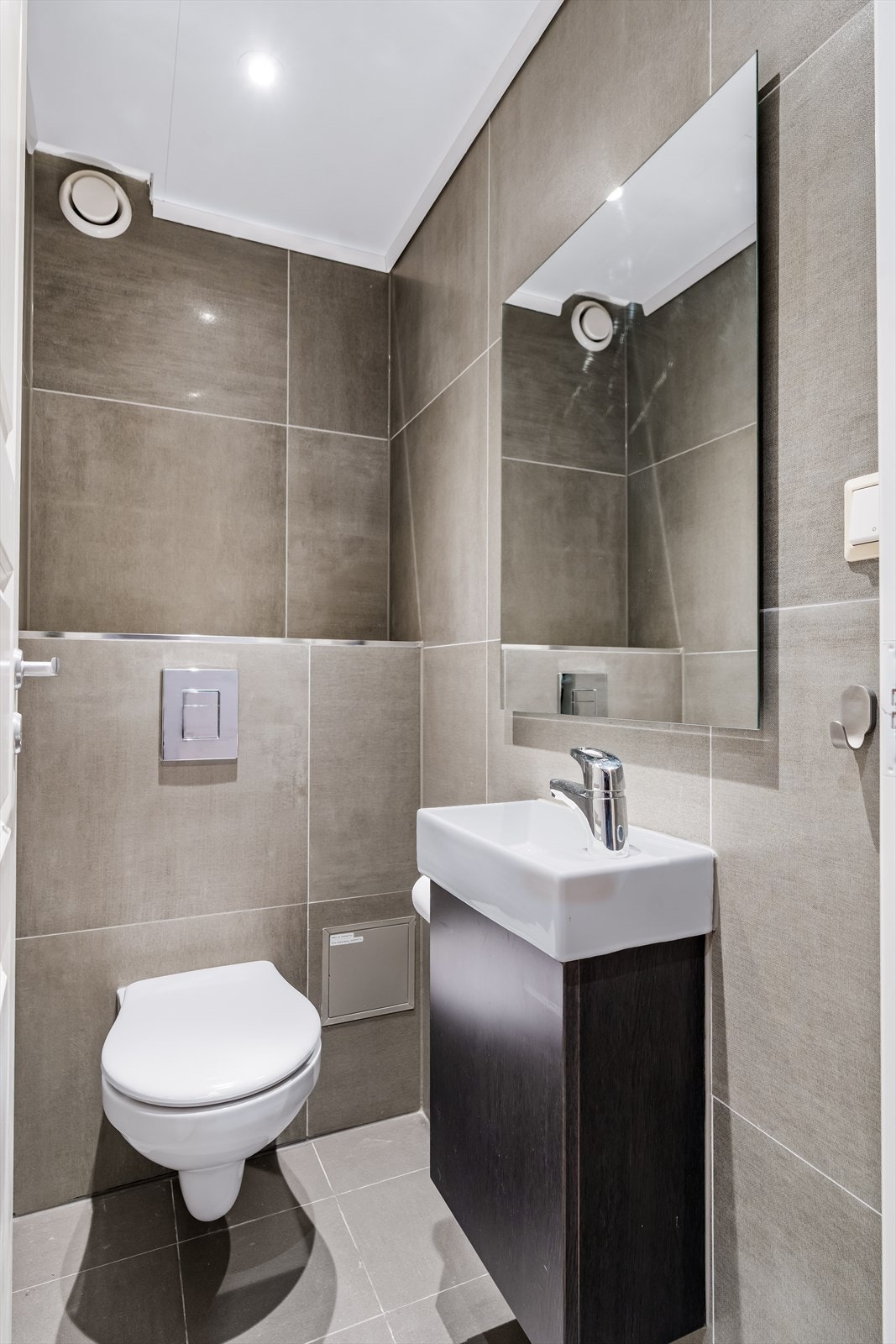 Vegg i vegg med badet ligger et separat toalettrom. Rommet er innredet med vegghengt toalett samt servant med underskap. Det er fliser på gulv og vegger. Galleribilde