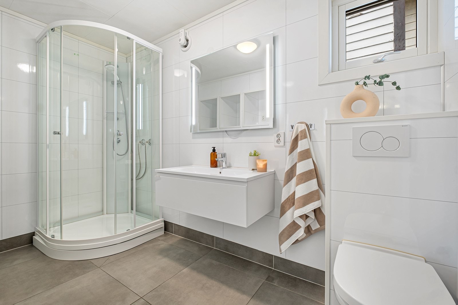 Badet er pent med flislagt gulv med varmekabler under og vegger med baderomsplater i flisimitasjon - Her finner du dusjkabinett, veggmontert toalett og baderomsinnredning Galleribilde