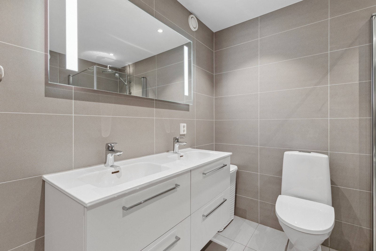 Smakfullt hovedbad plassert i egen suiteløsning med delikate fliser på gulv- og veggflater, samt varmekabler i gulv. Innredningen består av dobbelt servantmøblement med 4 skuffer, speil m/integrert belysning og toalett. Galleribilde