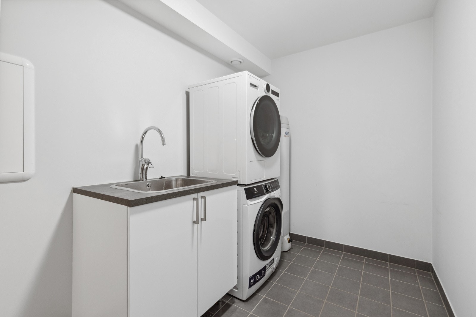 Familievennlig vaskerom med fliser på gulv med gulvvarme og malte slette veggflater. Inneholder bla. utslagsvask, benkeinnredning, varmtvannsbereder, samt opplegg for både vaskemaskin og tørketrommel i vaskesøyle. Galleribilde