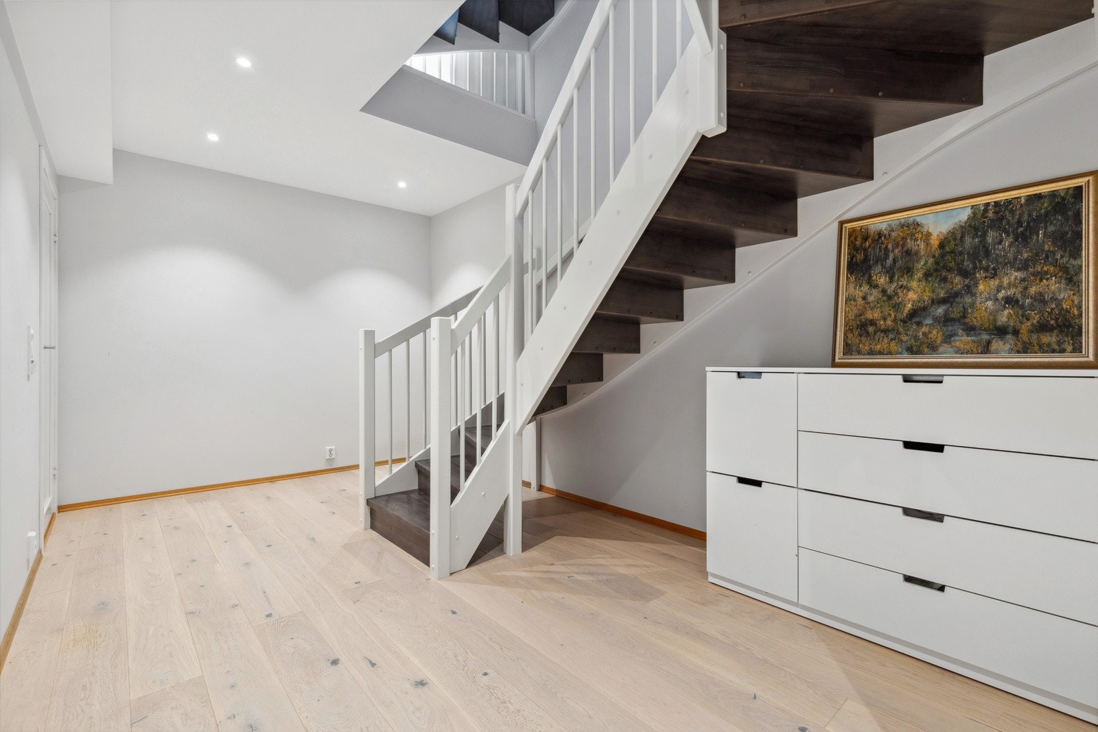 Stor og romslig gang i boligens sokkeletasje med lyspigmentert parkett på gulv og downlights i himling. Galleribilde