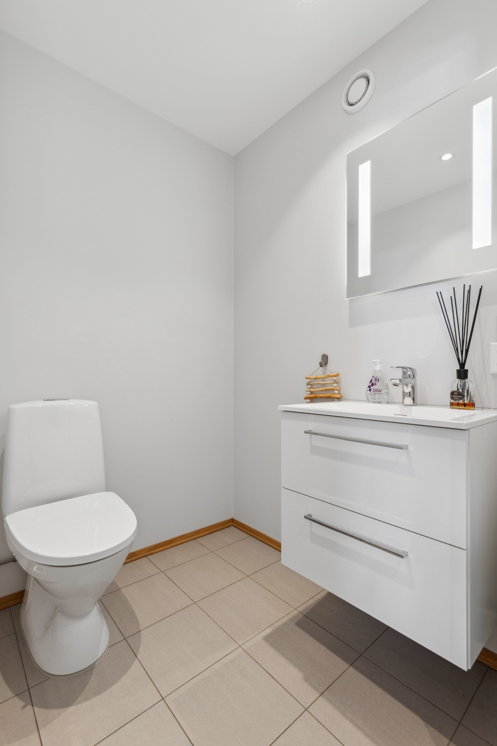 Praktisk gjestetoalett med fliser på gulv med gulvvarme og malte slette veggflater. Inneholder bla. servantinnredning, speil og toalett. Galleribilde