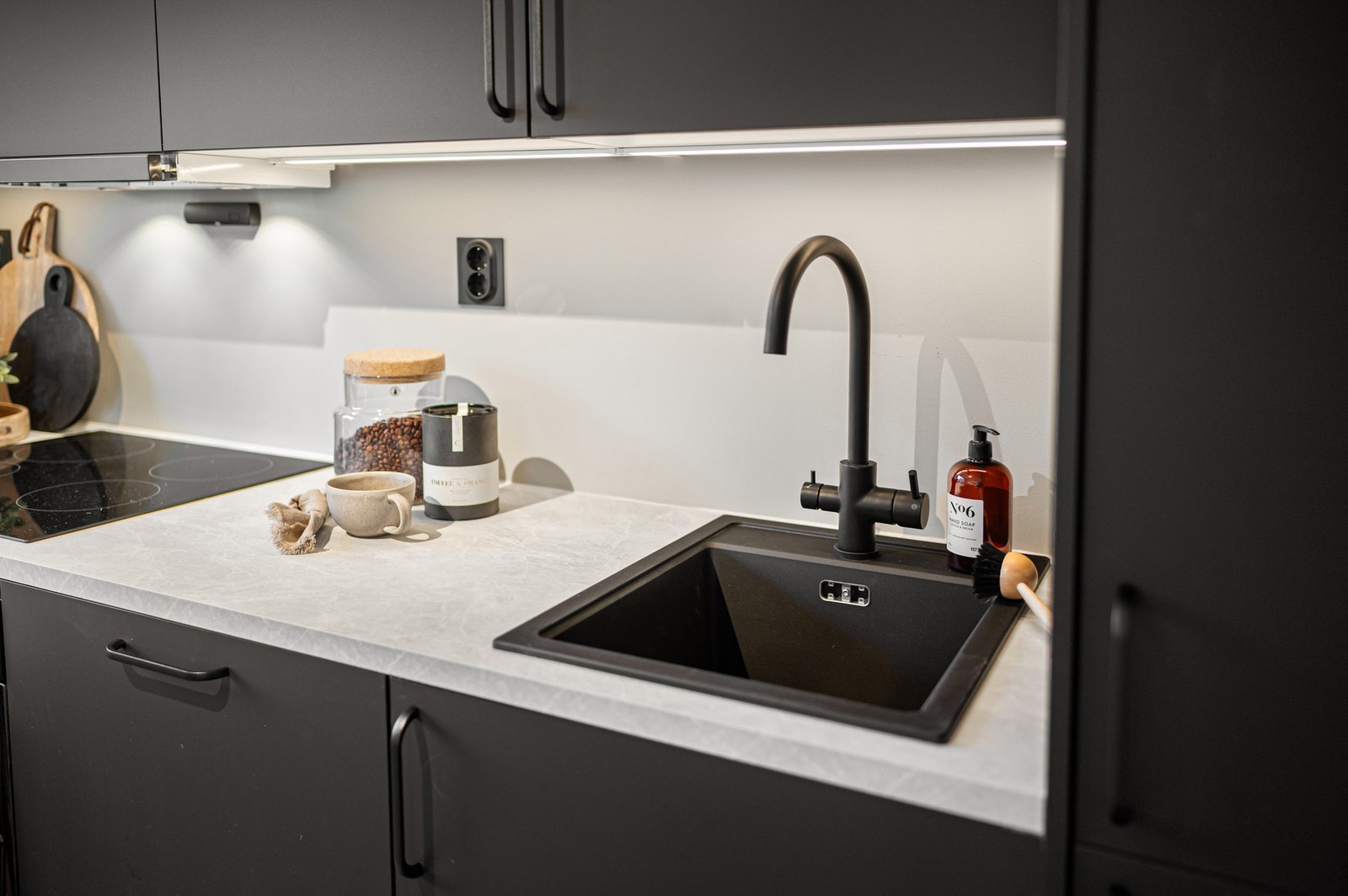 Legg merke til de gjennomgående sorte detaljene i form av fronter, håndtak, vask, kjøkkenarmatur, stikkontakter og komfyr. Den lyse benkeplaten gir kjøkkenet en naturlig og fin kontrast. Galleribilde