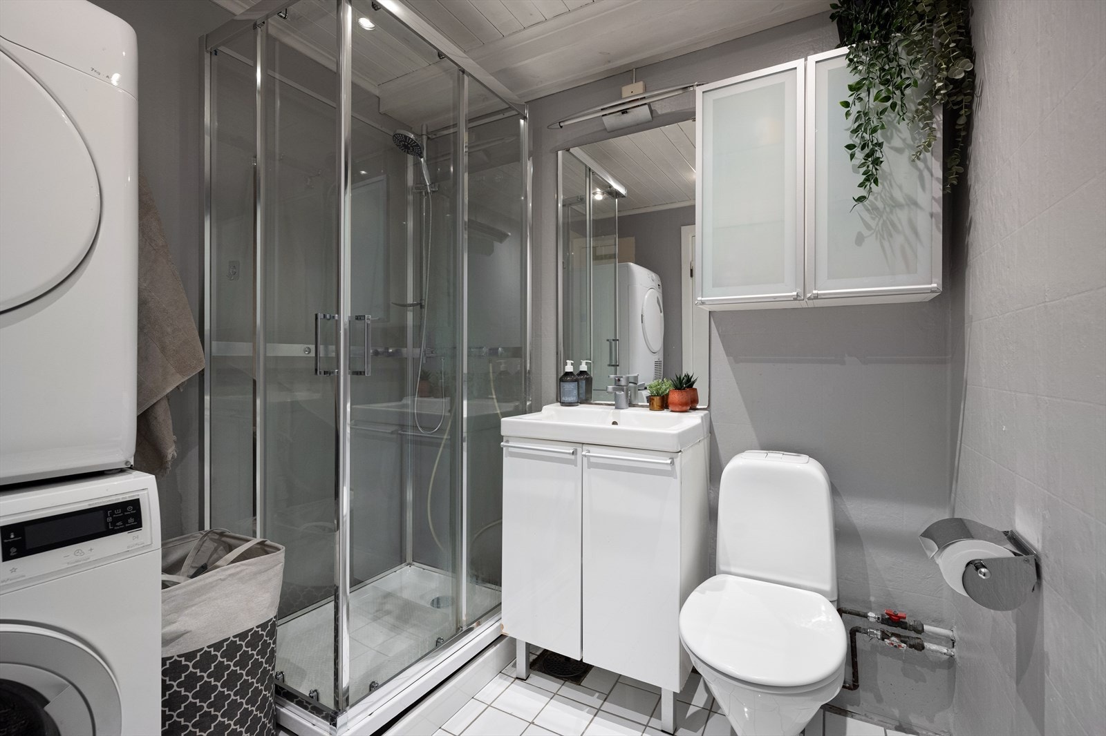 Badet er holdt i grått og hvitt og er innredet med toalett, dusjkabinett, vask i skapinnredning og opplegg for vaskemaskin og tørketrommel. Galleribilde