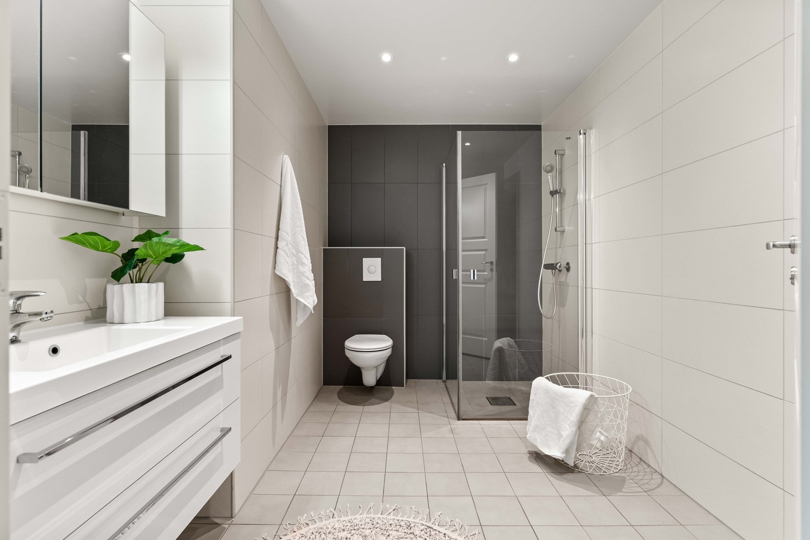 Stilrent, flislagt baderom med moderne innredning. Badet er utstyrt med vegghengt toalett, romslig servantmøbel med gode skuffer og lekkert dusjhjørne med innfellbare glassdører. Downlights i taket og nøytrale fargevalg skaper en behagelig atmosfære. Galleribilde