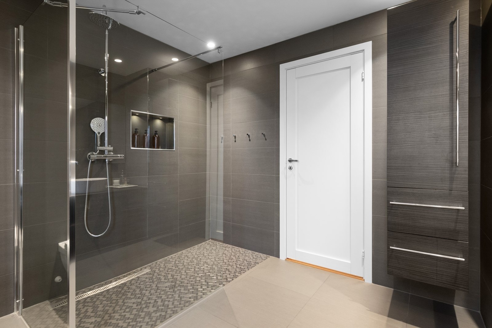 Fliser på gulv og vegger, baderom inneholder dusjhjørne, veggmontert klosett og servant i innredning. Galleribilde