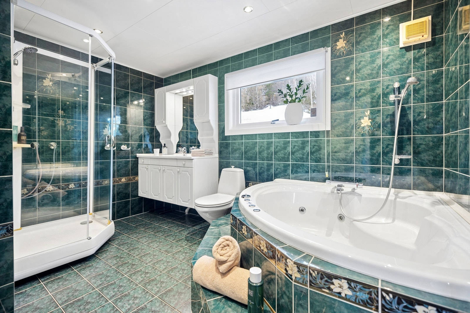 Romslig flislagt bad med gulvvarme. Vegghengt baderomsinnredning med profilerte lyse fronter, dusjkabinett, hjørnebadekar og gulvstående toalett. Galleribilde