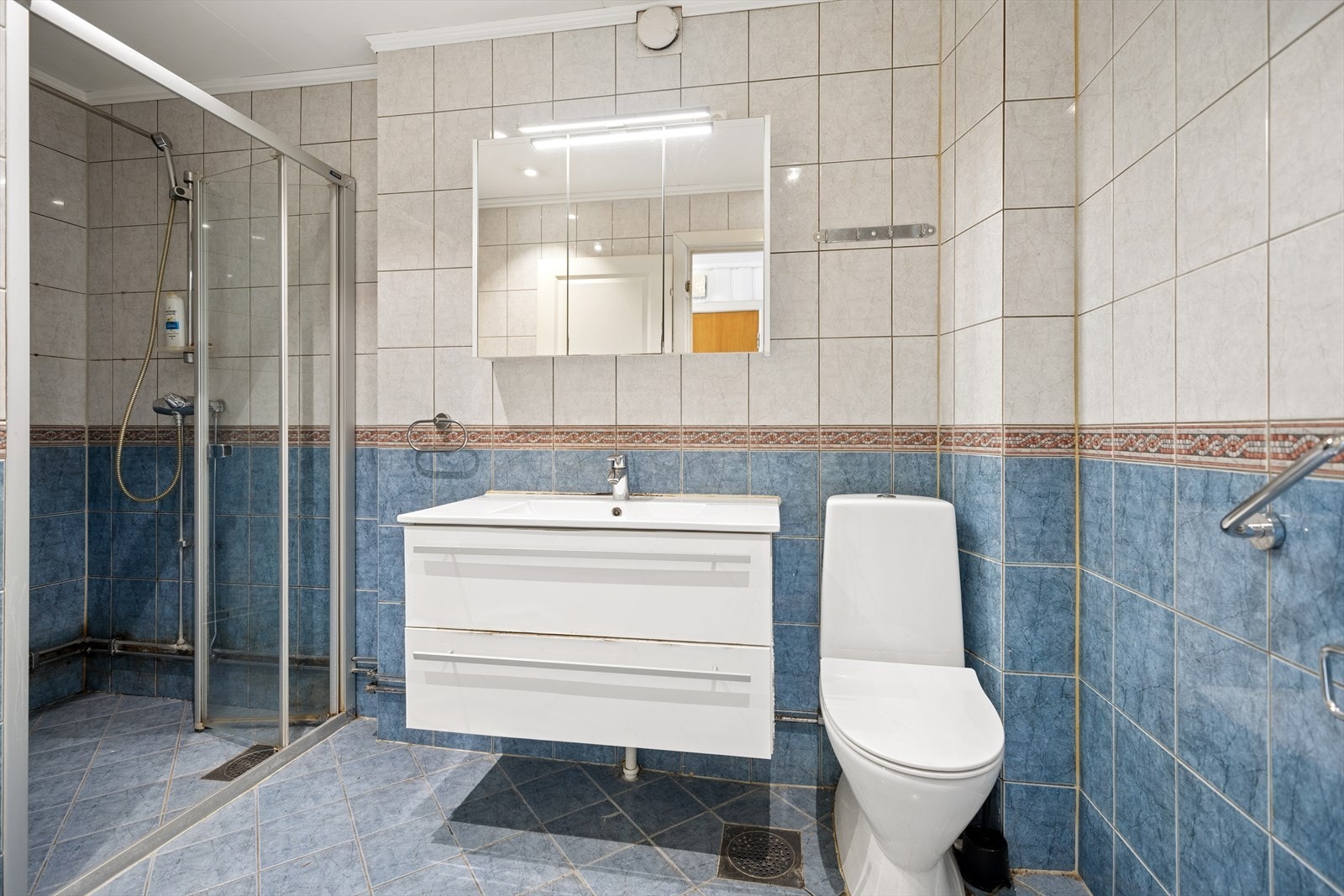 Badet er flislagt på vegger og gulv, med elektriske varmekabler i gulvet. Rommet har baderomsinnredning med nedfelt servant, overskap med speil og lys, toalett og dusjhjørne Galleribilde