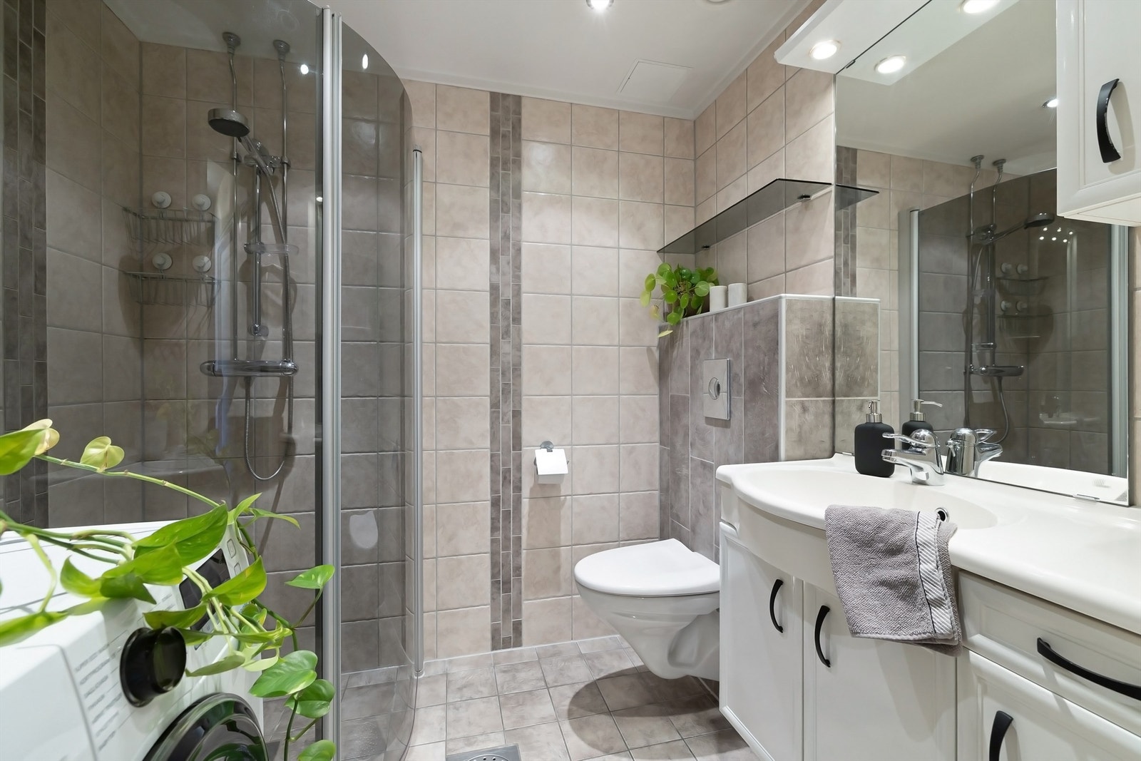 Delikat flislagt bad med gulvvarme og downlights utstyrt med vegghengt toalett, dusjhjørne med praktiske, innfellbare glassdører og opplegg for vaskemaskin Galleribilde