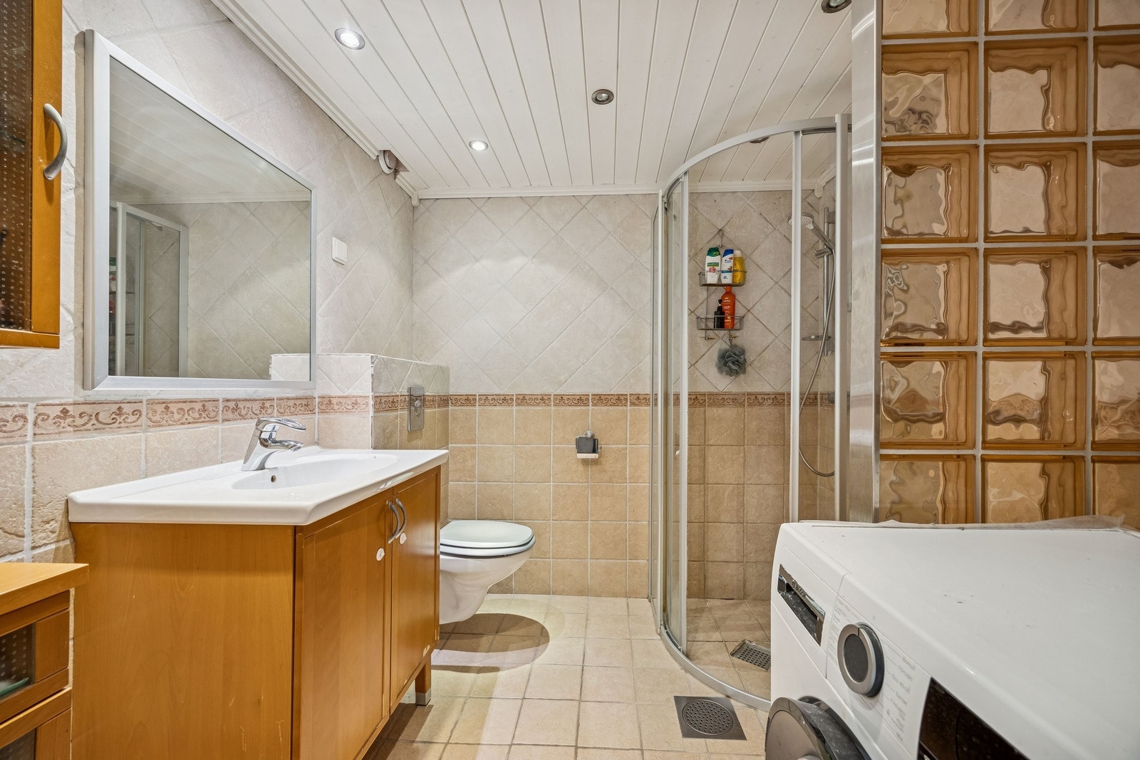 Delikat bad med bestående av servant, skapinnredning, dusjsone med innfellbare dører og opplegg for vaskemaskin Galleribilde