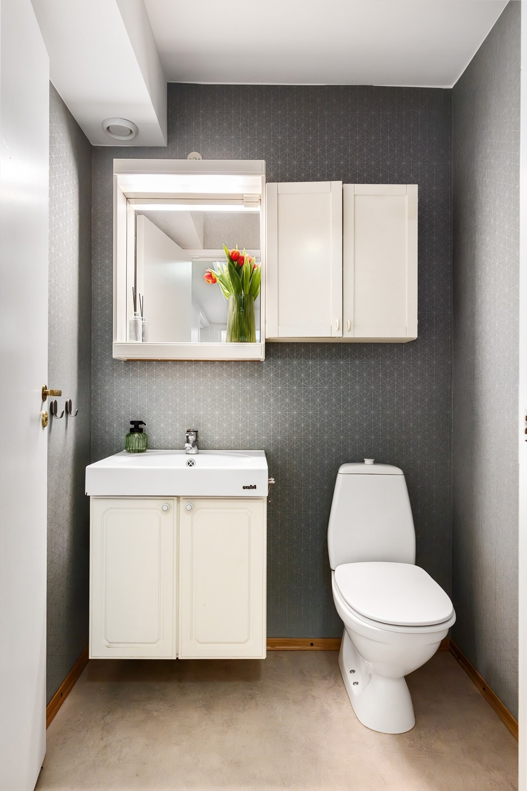 Praktisk toalettrom med belegg på gulv, tapet på vegger og malt himling. Rommet består av gulvmontert klosett, servantskap med nedfelt servant, veggskap og speil med lys over. Galleribilde