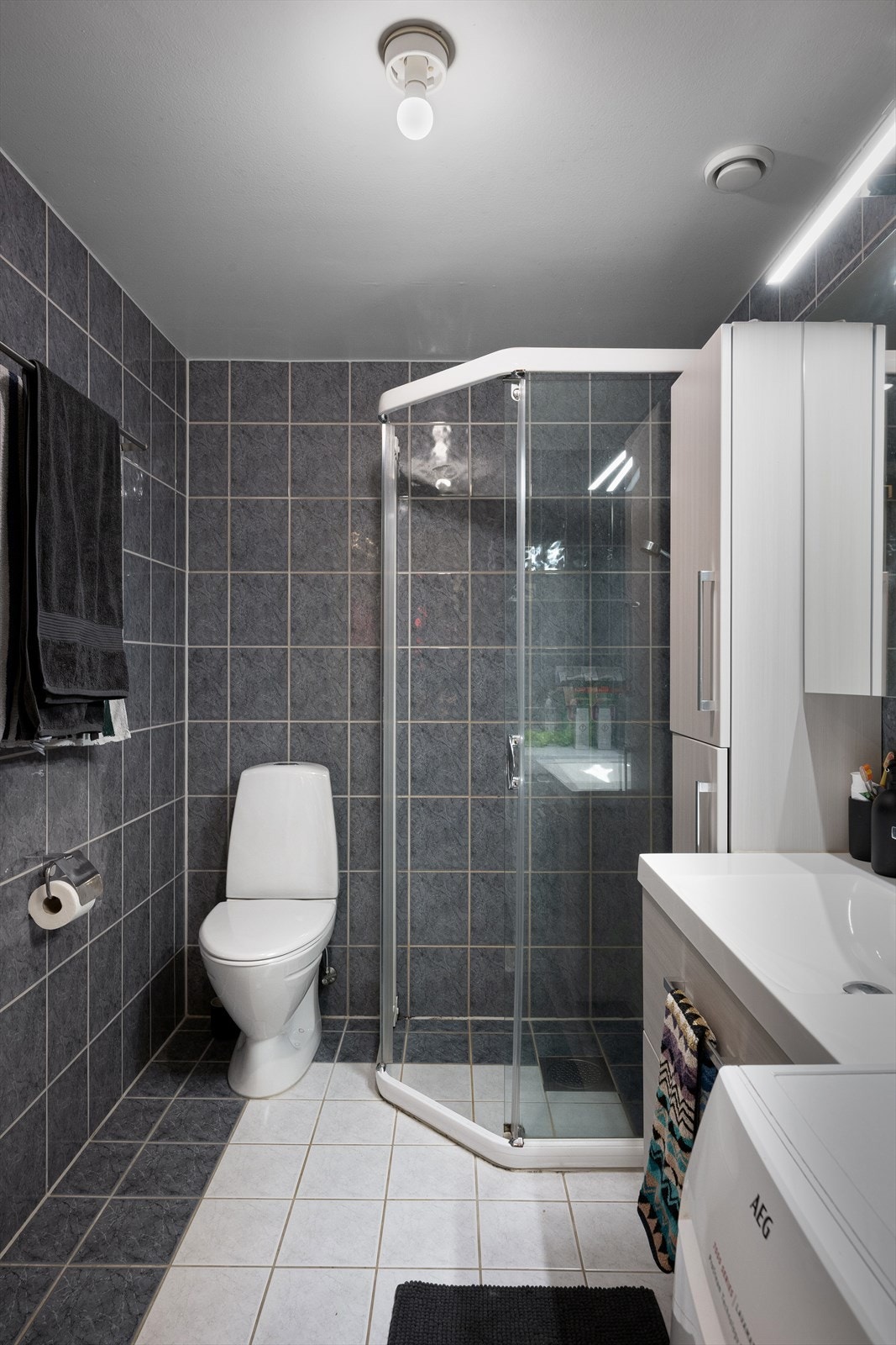 Flott bad med fliser på vegg og gulv. Badet er utstyrt med gulvmontert toalett, dusjhjørne med glassdør i klart glass, lys baderomsinnredning med servant og speil. Opplegg for vaskemaskin. Vannbåren varme i gulv. Galleribilde