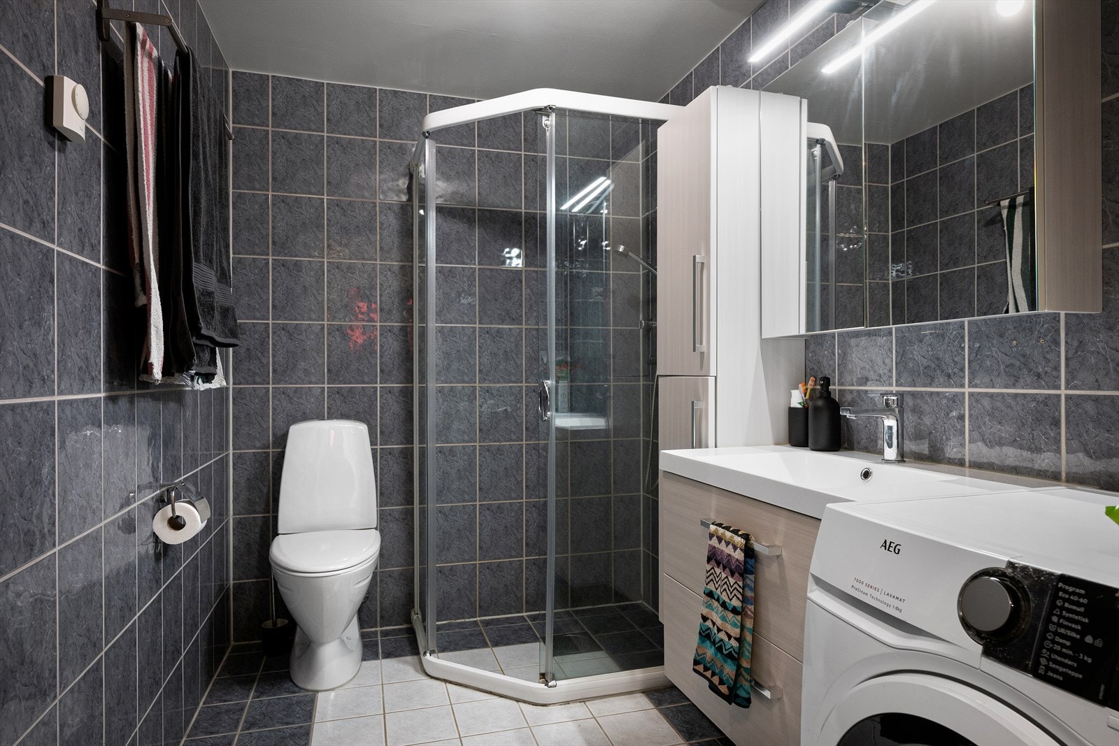 Flott bad med fliser på vegg og gulv. Badet er utstyrt med gulvmontert toalett, dusjhjørne med glassdør i klart glass, lys baderomsinnredning med servant og speil. Opplegg for vaskemaskin. Vannbåren varme i gulv. Galleribilde