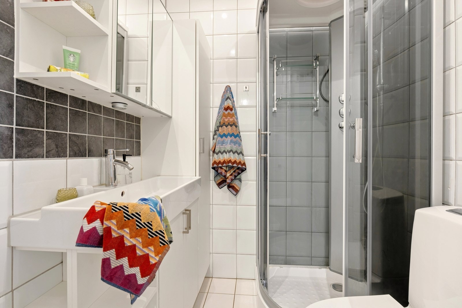 Badet har flislagte overflater på gulv og vegger. Det er elektriske varmekabler i gulvet, og belysningen via innfelte downlights i taket. Innredningen består av en servantseksjon med nedfelt servant, toalett og et dusjkabinet. Galleribilde