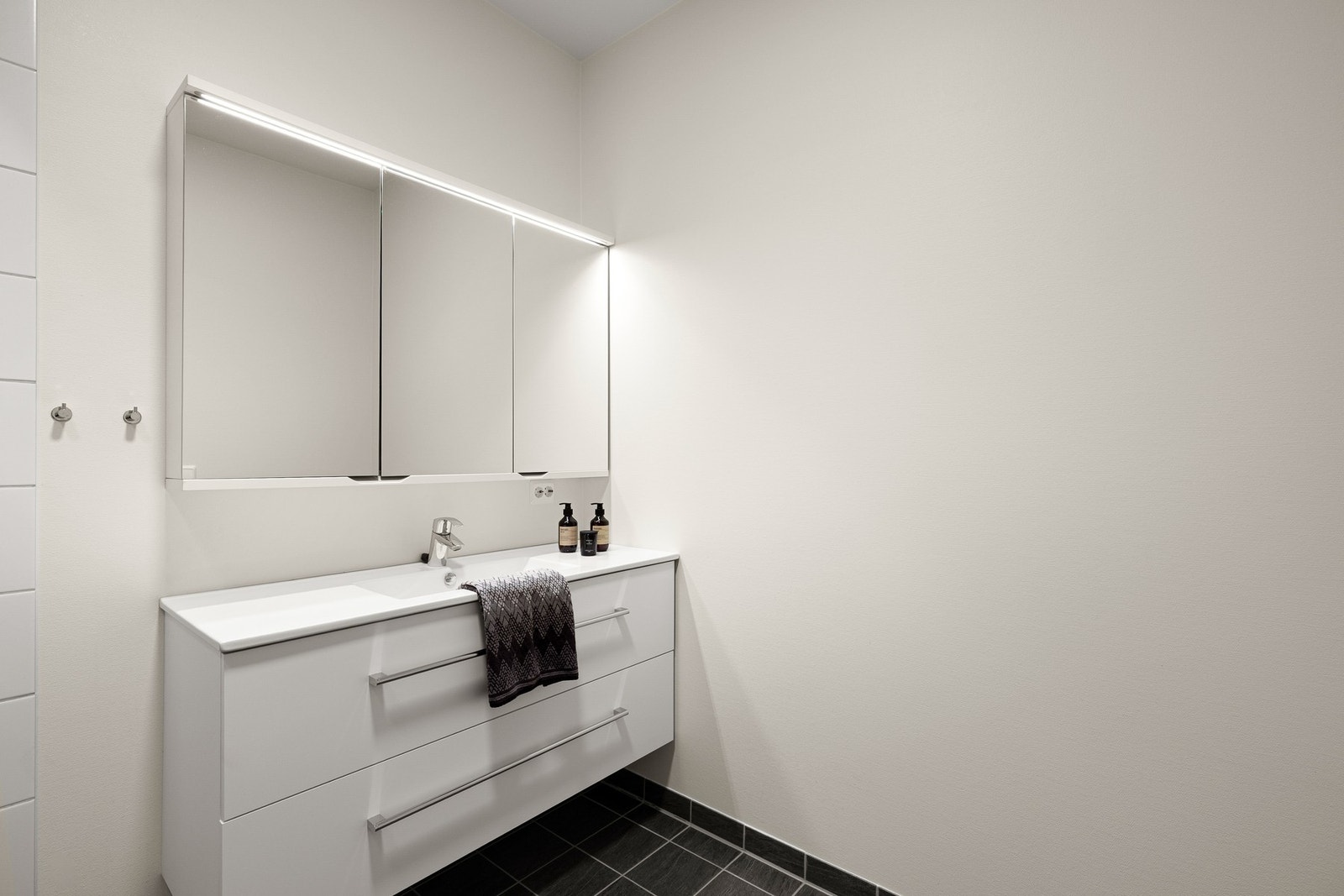 Baderomsinnredningen har hvite, glatte fronter og består av en heldekkende servant på en skuffeseksjon, et stort speil med god belysning. Galleribilde