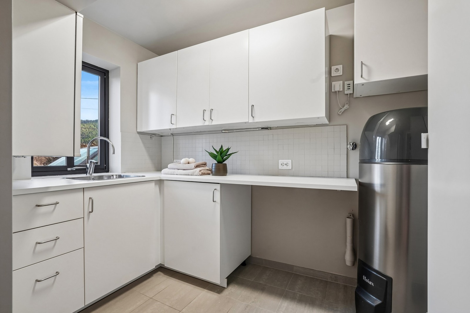 Praktisk vaskerom med flislagt gulv og elektriske varmekabler. Innredet med opplegg for vaskemaskin, innredning med hvite glatte fronter, overskap og laminat benkeplate med nedfelt stålkum. Galleribilde