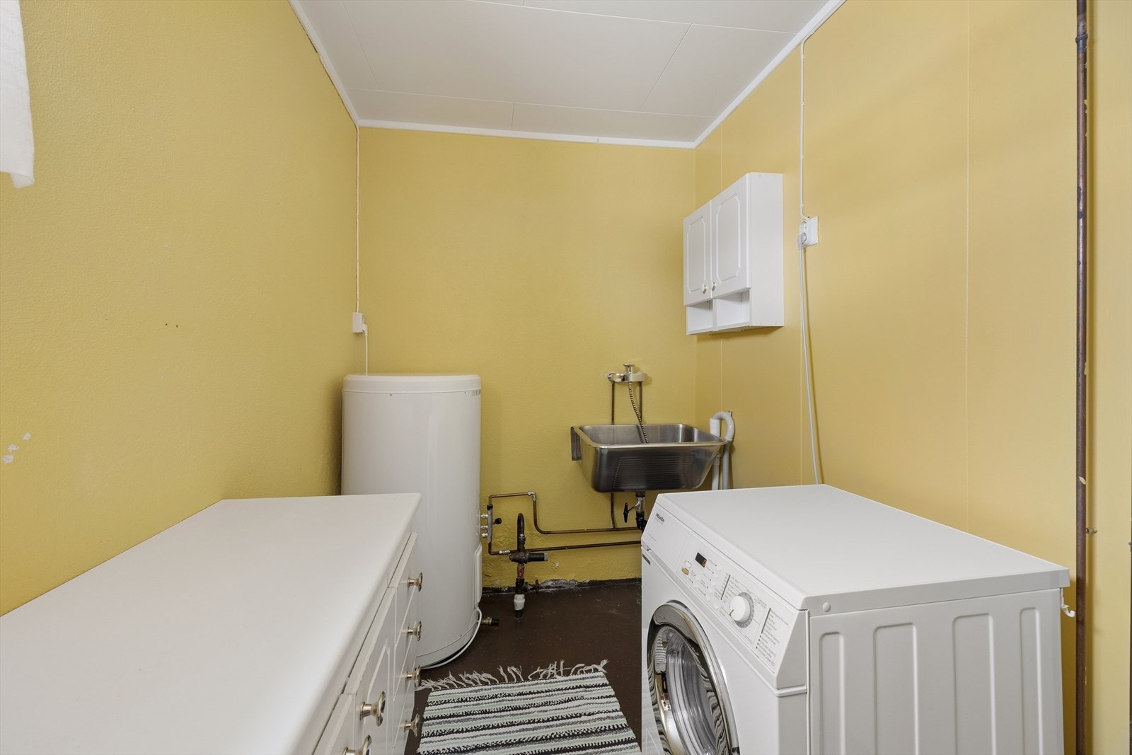 Praktisk vaskerom med malt betonggulv og opplegg for vaskemaskin. Varmtvannstanken er på ca. 200 liter. Galleribilde