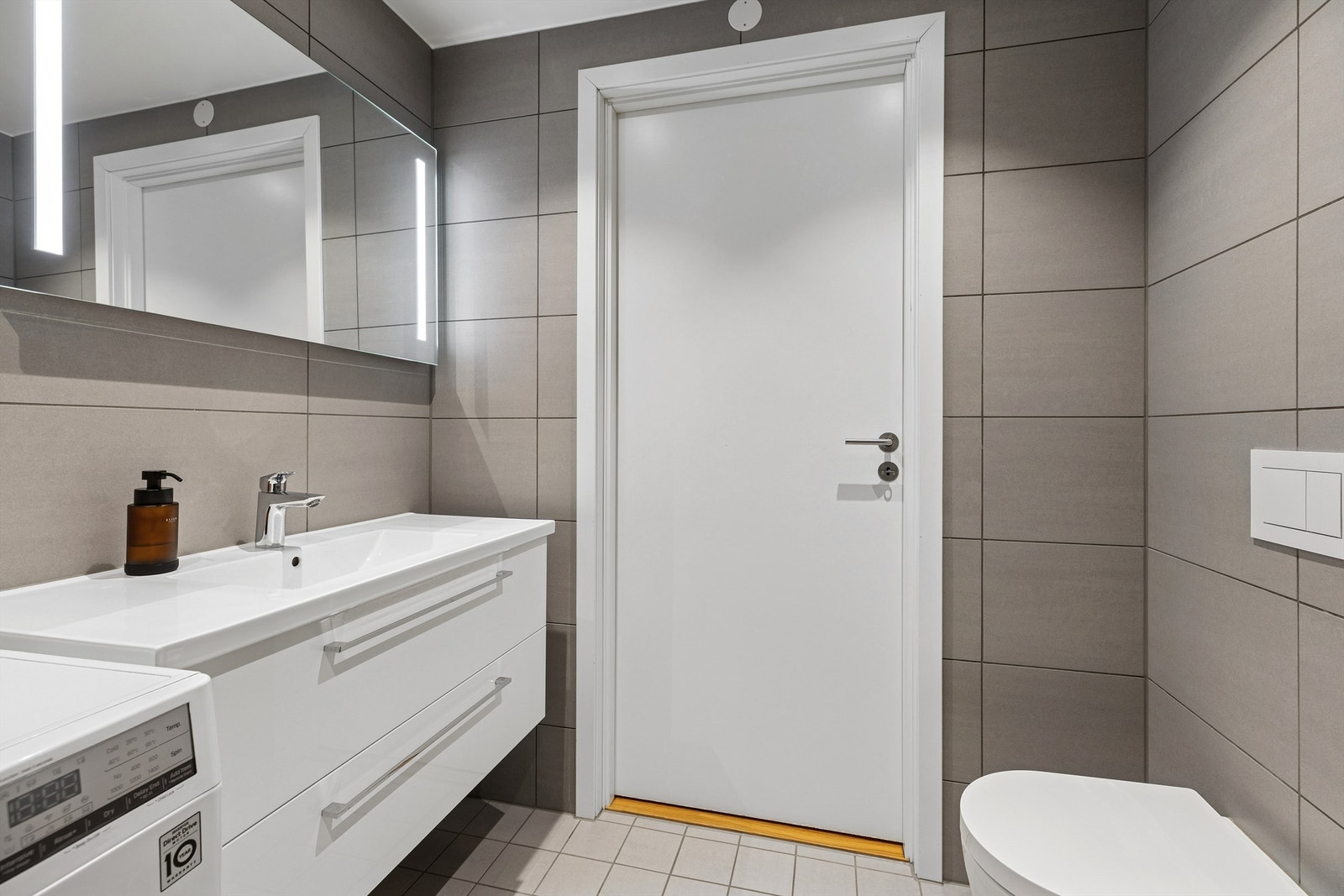 Lekkert flislagt bad med gulvvarme. Her finnes dusjhjørne med innfellbare glassdører, moderne servantinnredning, vegghengt toalett og opplegg for vaskemaskin. Galleribilde