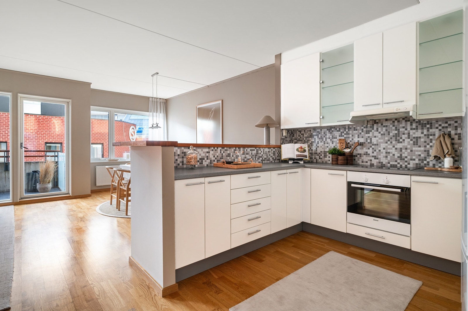 Kjøkkeninnredningen har glatte fronter, laminat benkeplate med nedfelt vaskekum, overskap og mosaikkfliser på vegg over benkeplaten. Galleribilde